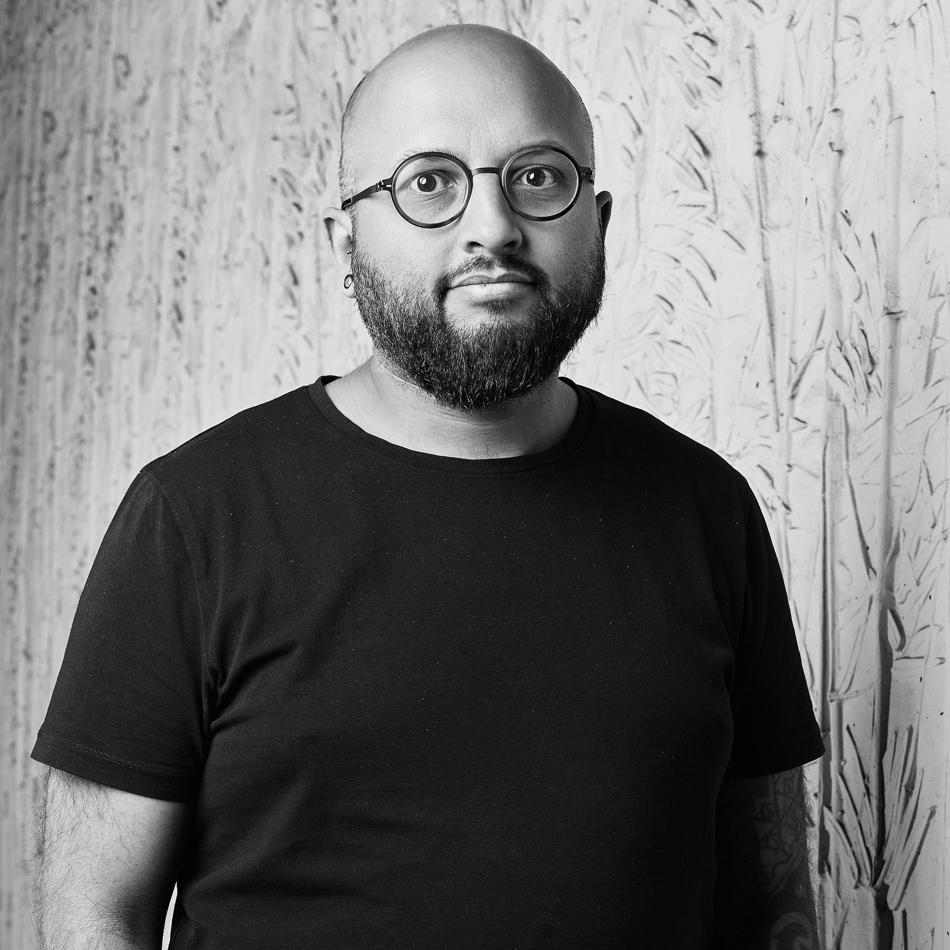 Portrait d'un homme à lunettes et à barbe portant un t-shirt noir devant un mur texturé