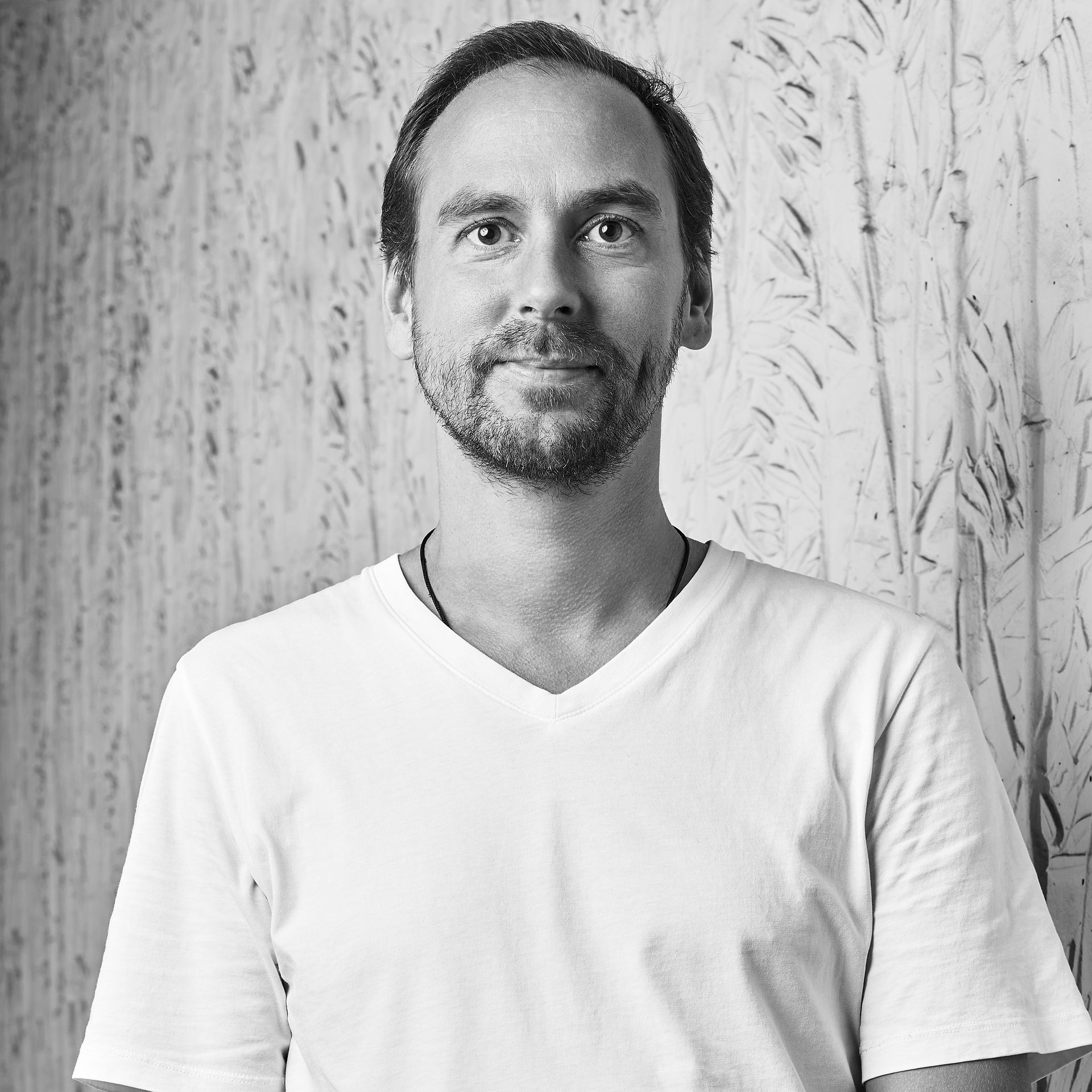 Portrait en noir et blanc d'une personne vêtue d'un t-shirt blanc à col en V, debout devant un mur texturé.