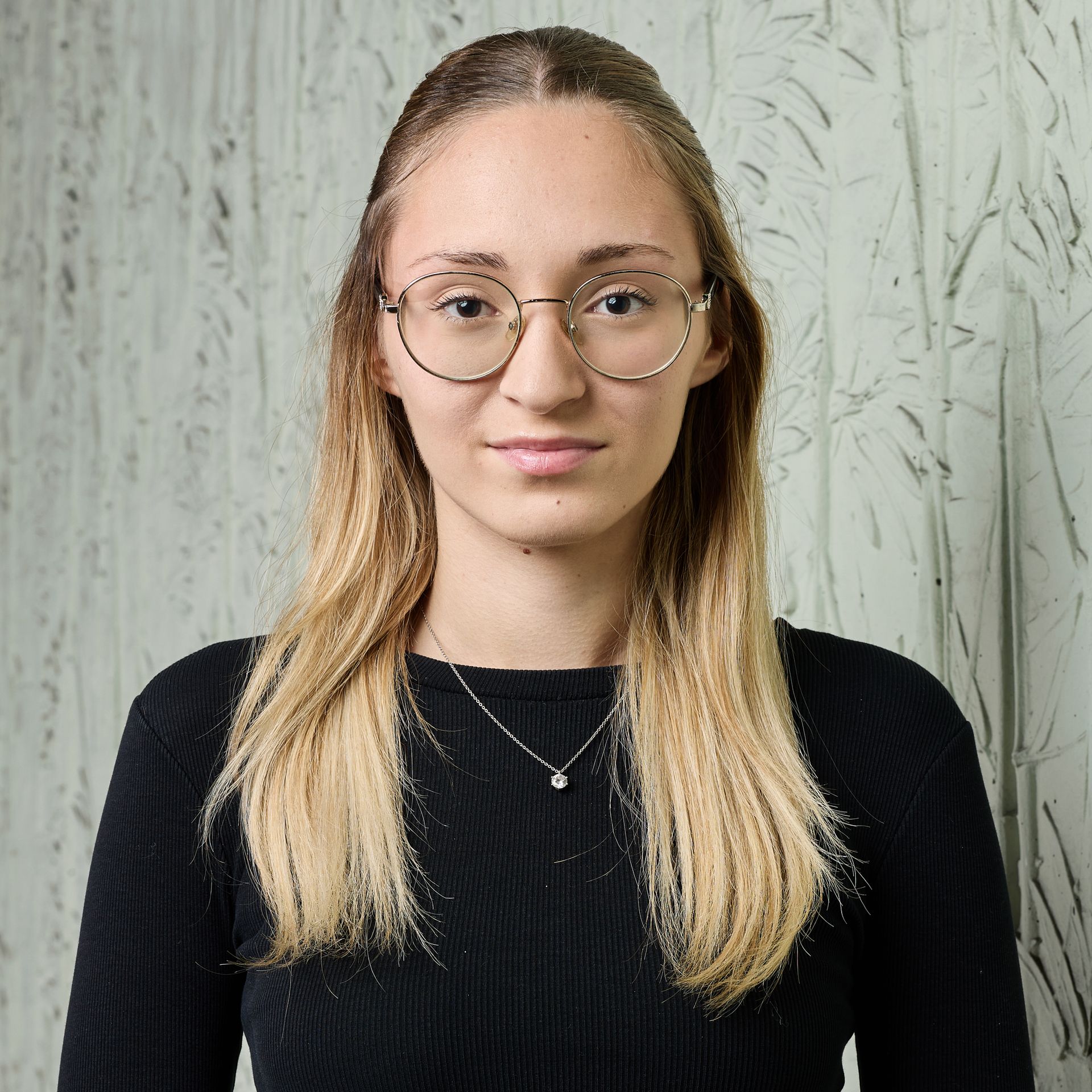 Portrait d'une personne portant des lunettes et un haut noir sur un fond légèrement texturé.
