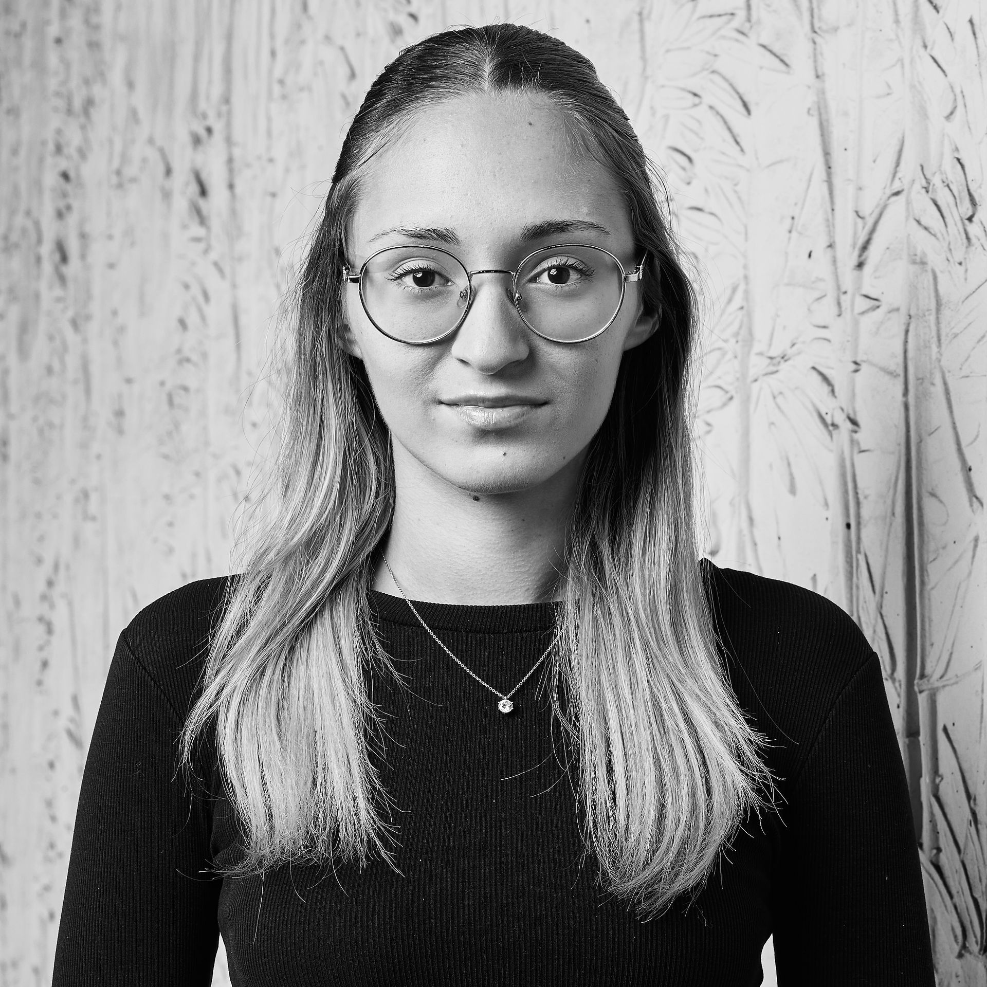 Portrait d'une personne portant des lunettes et un haut foncé, se détachant sur un fond clair à motifs.