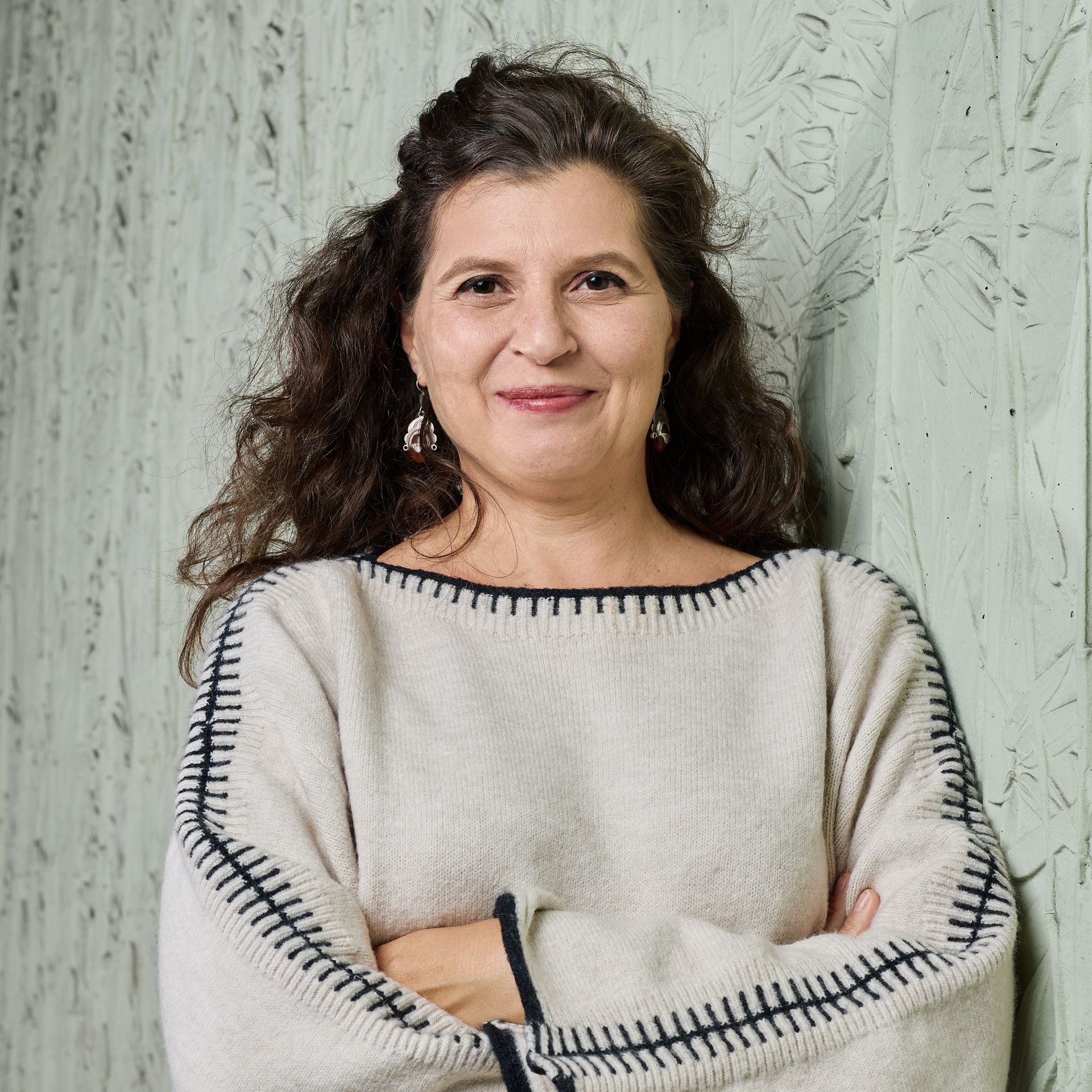 Femme souriante en pull blanc, les bras croisés, contre un mur à la texture claire