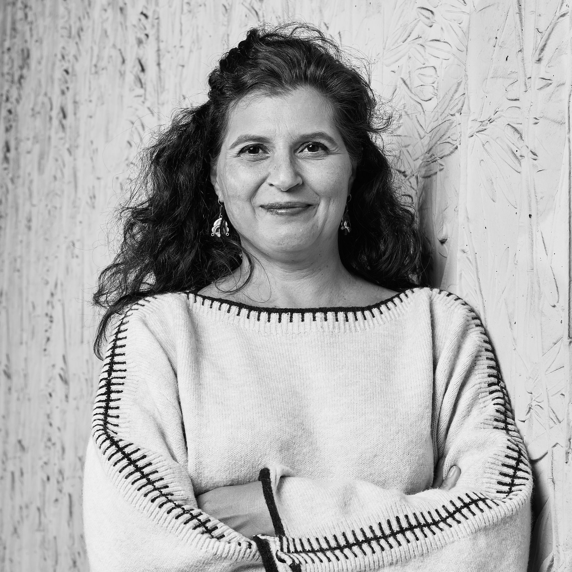 Portrait d'une personne souriante, les bras croisés, portant un pull léger, devant un mur texturé.