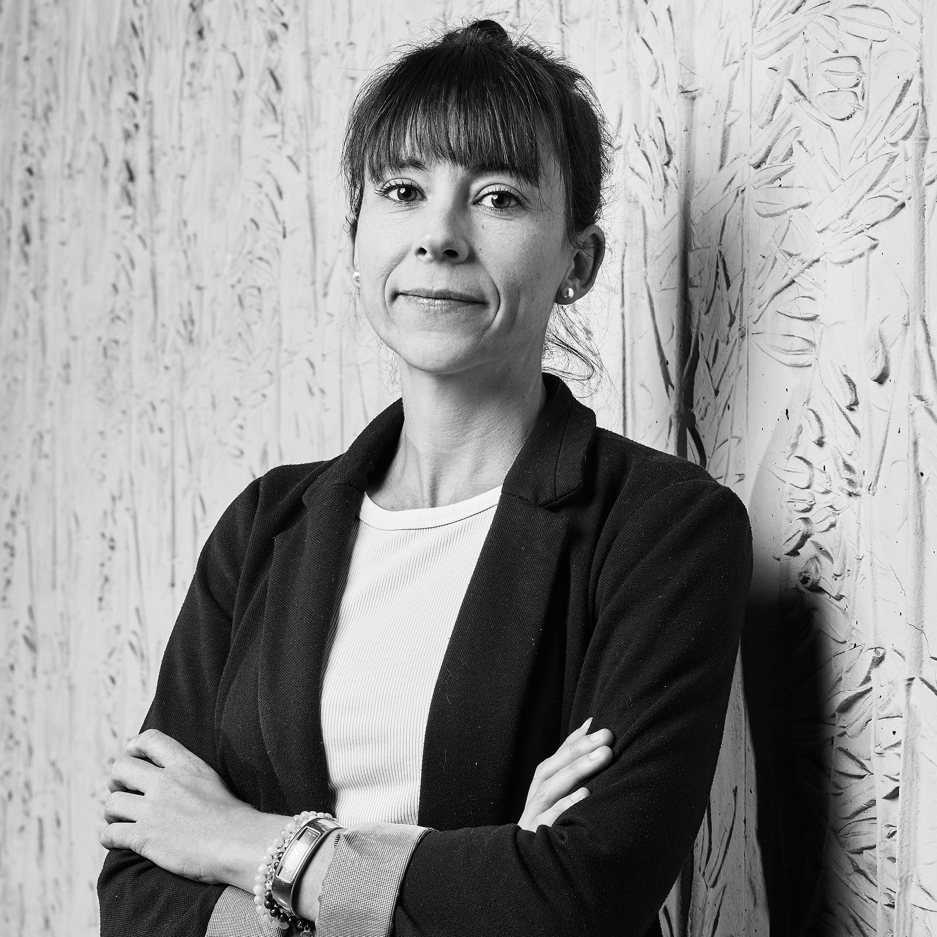Personne debout, les bras croisés, contre un mur texturé, portant un blazer foncé et un haut blanc.