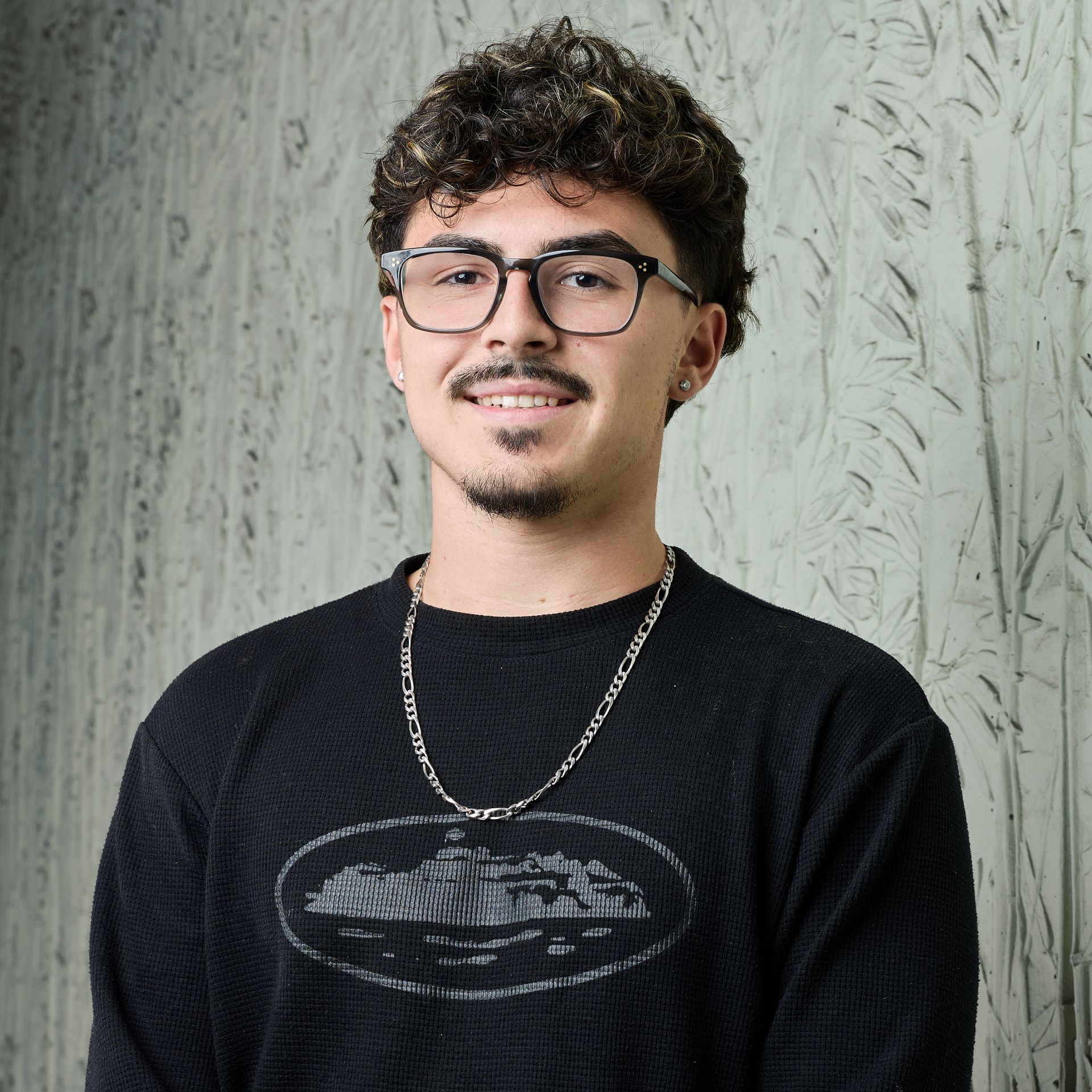 Personne souriante portant des lunettes, des cheveux bouclés, une chemise noire et un collier à chaînes, devant un mur texturé.