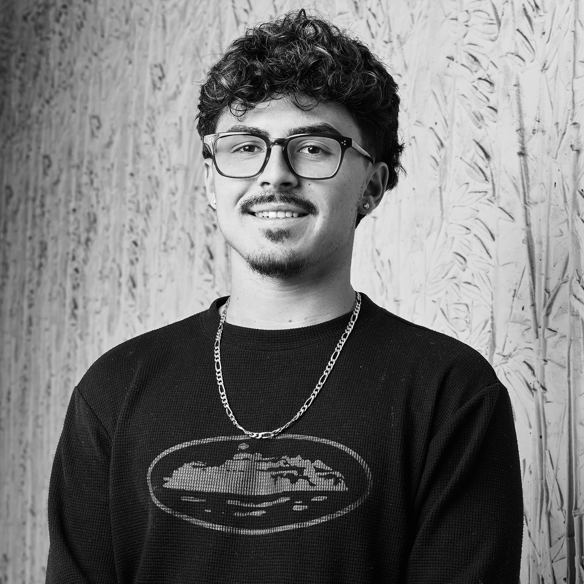 Personne aux cheveux bouclés et portant des lunettes, un pull foncé et une chaîne, debout contre un mur texturé.