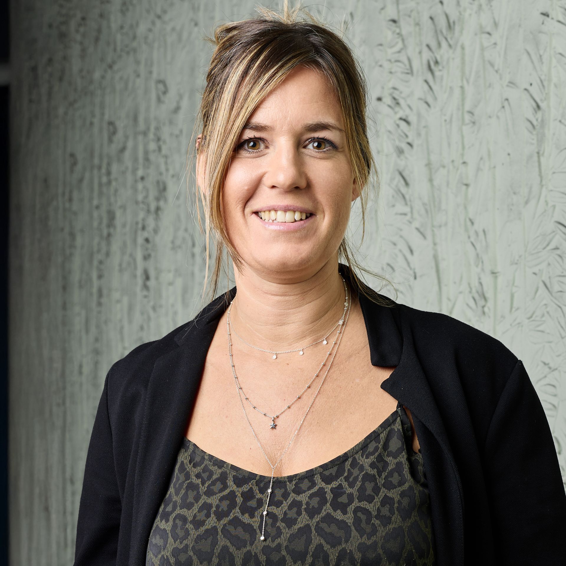 Femme souriante vêtue d'un cardigan noir et d'un haut à motifs, debout devant un mur gris texturé.
