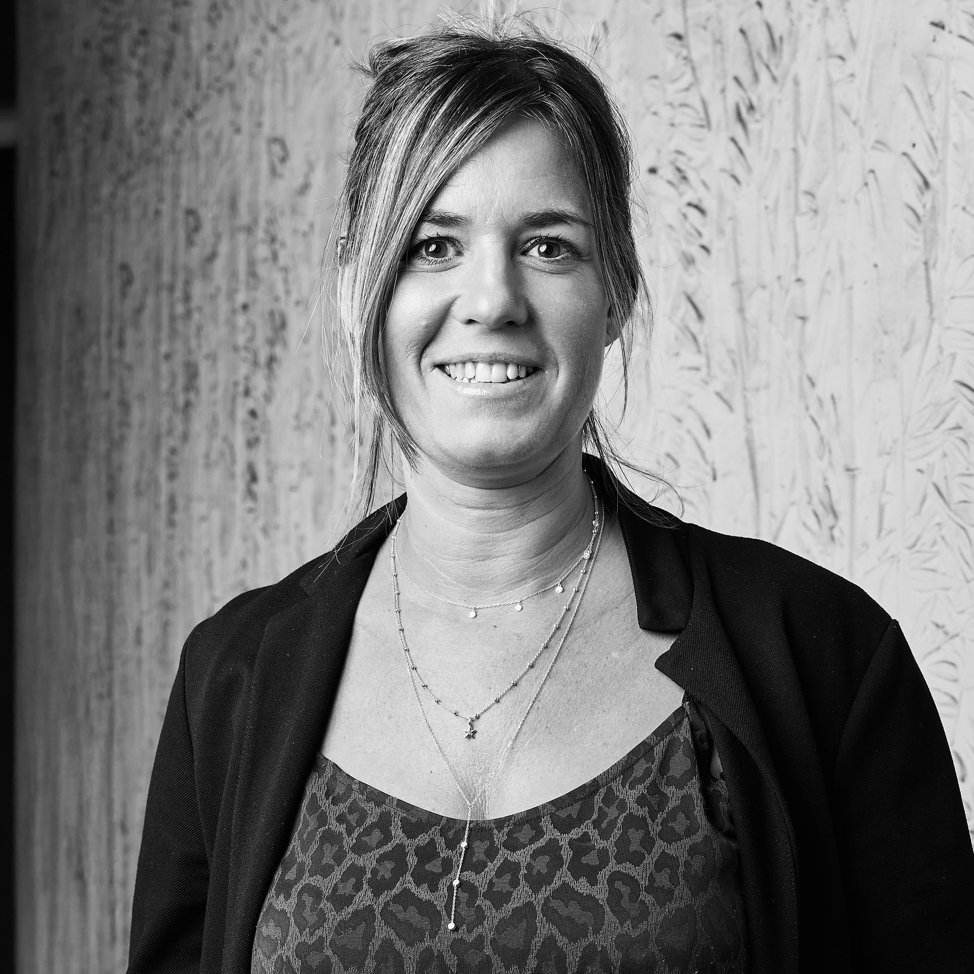 Femme souriante vêtue d'un cardigan foncé et d'un haut à motifs, debout contre un mur texturé