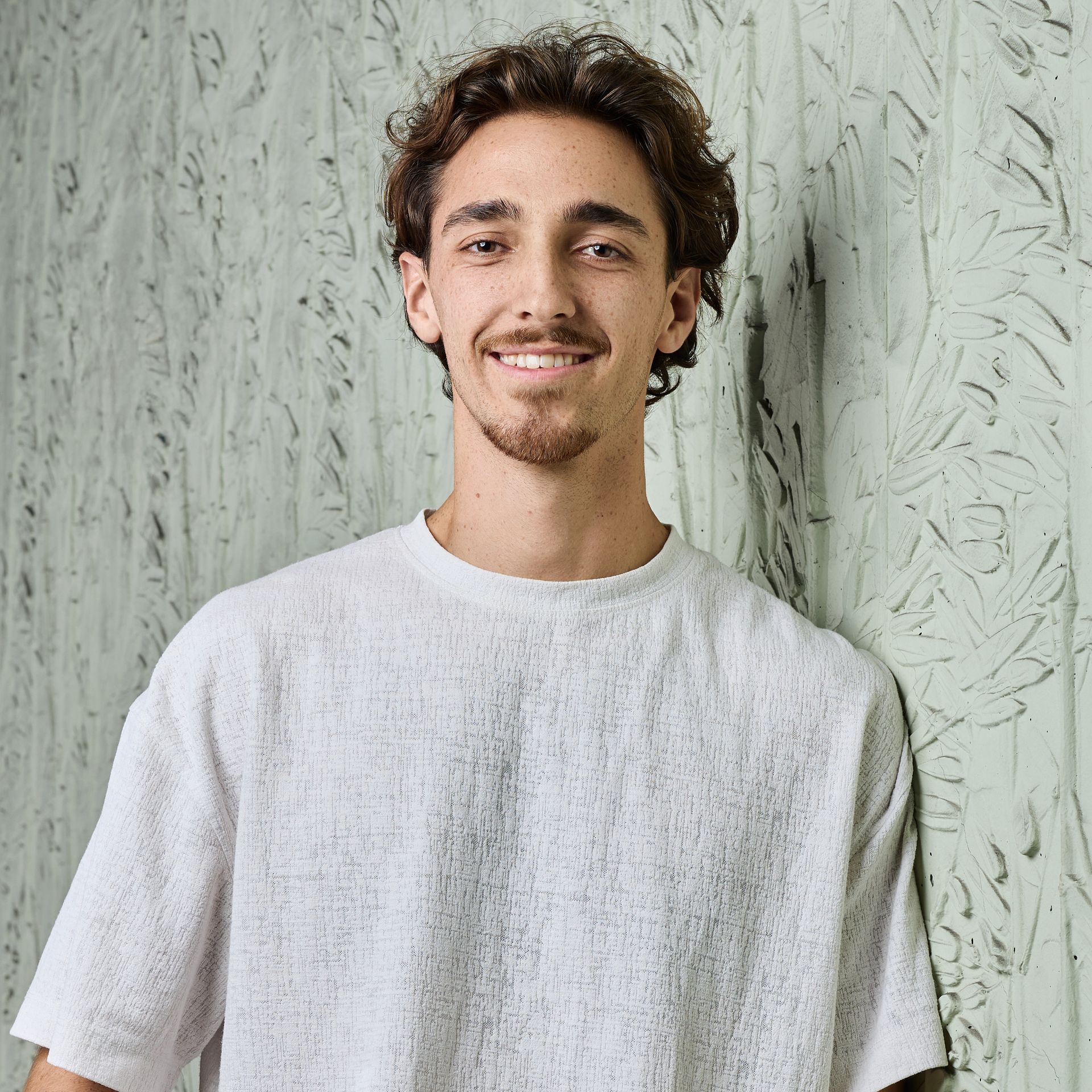 Un homme souriant, vêtu d'une chemise blanche texturée, se tient devant un mur texturé gris clair.