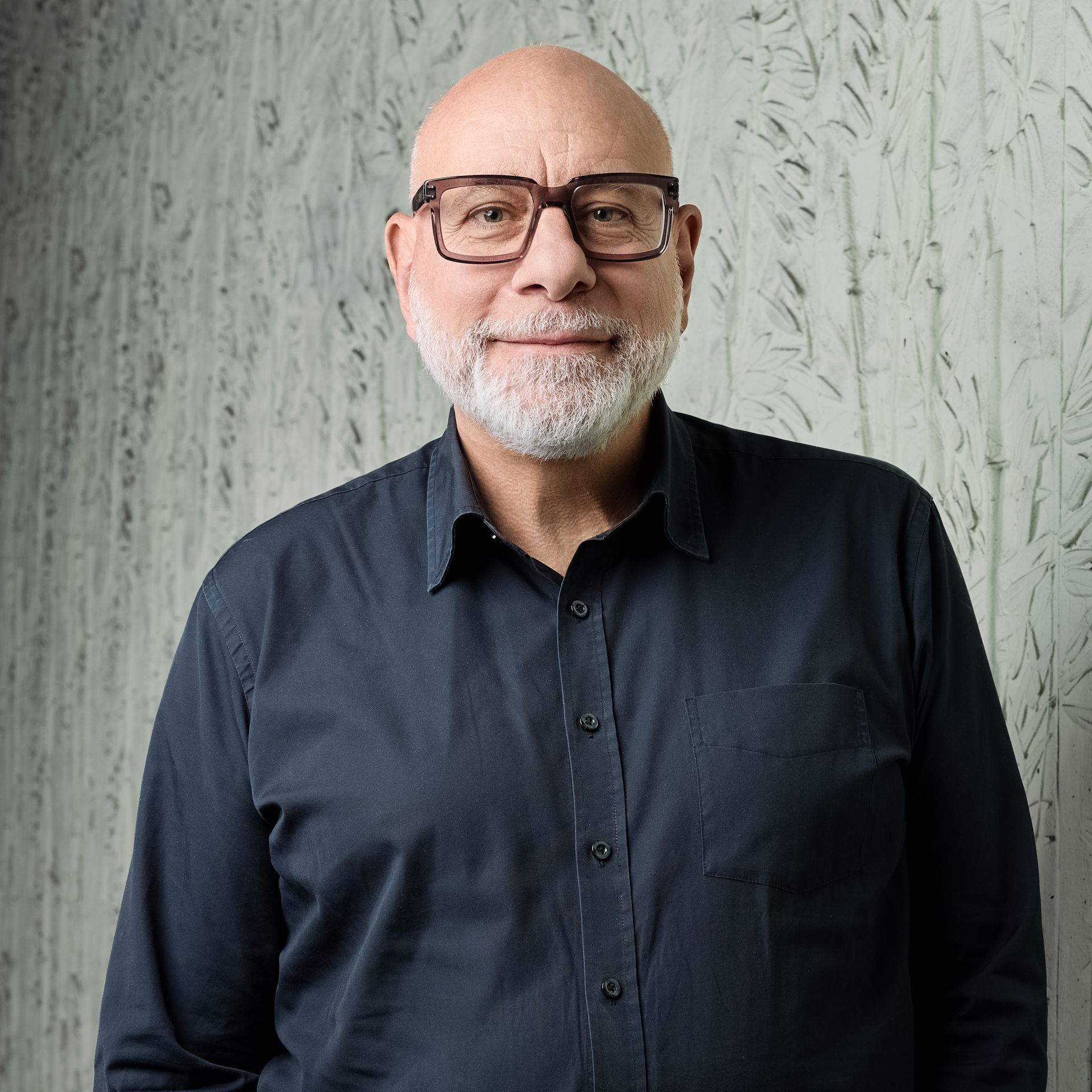 Un homme chauve souriant, portant des lunettes et une barbe grise, vêtu d'une chemise sombre, se détache sur un mur gris texturé.