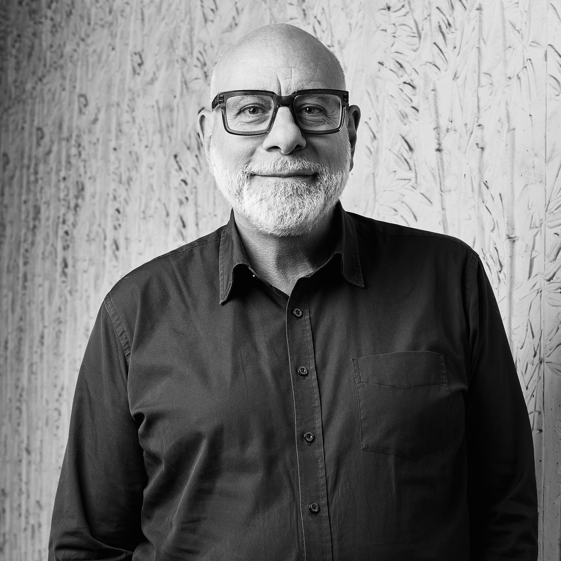Un homme souriant, portant des lunettes et une chemise sombre, se tient devant un mur texturé en noir et blanc.