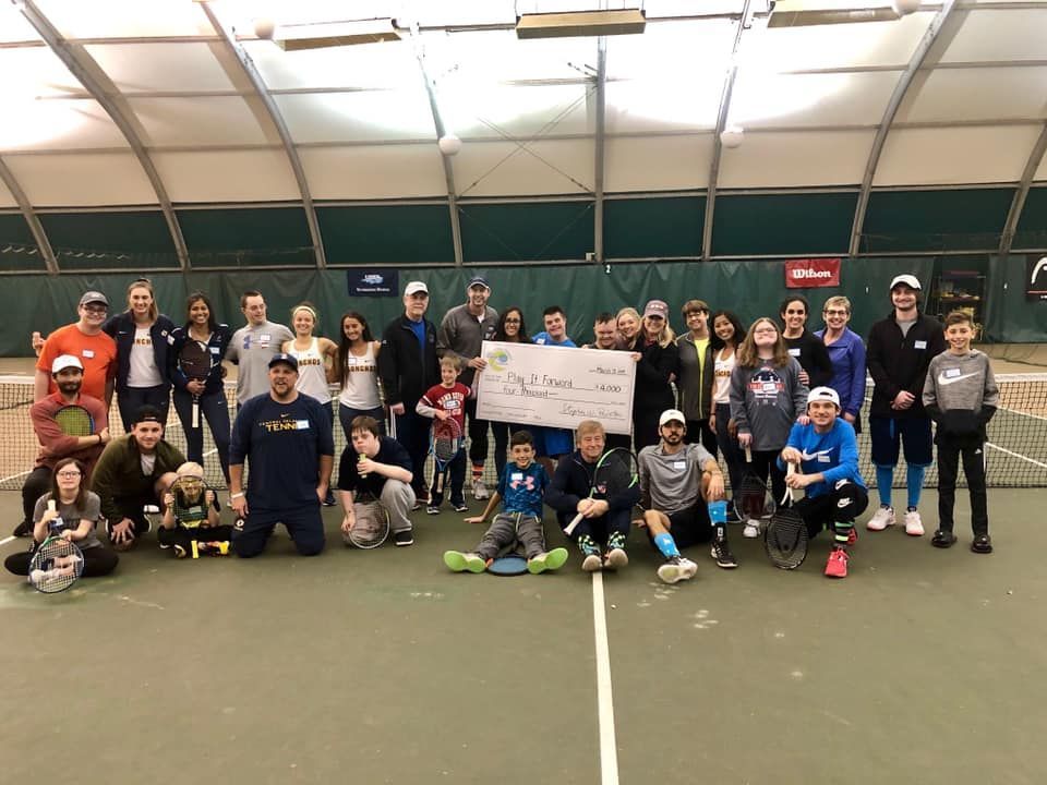 Group of people posing indoors on a tennis court, holding a large sign and wearing sports gear.