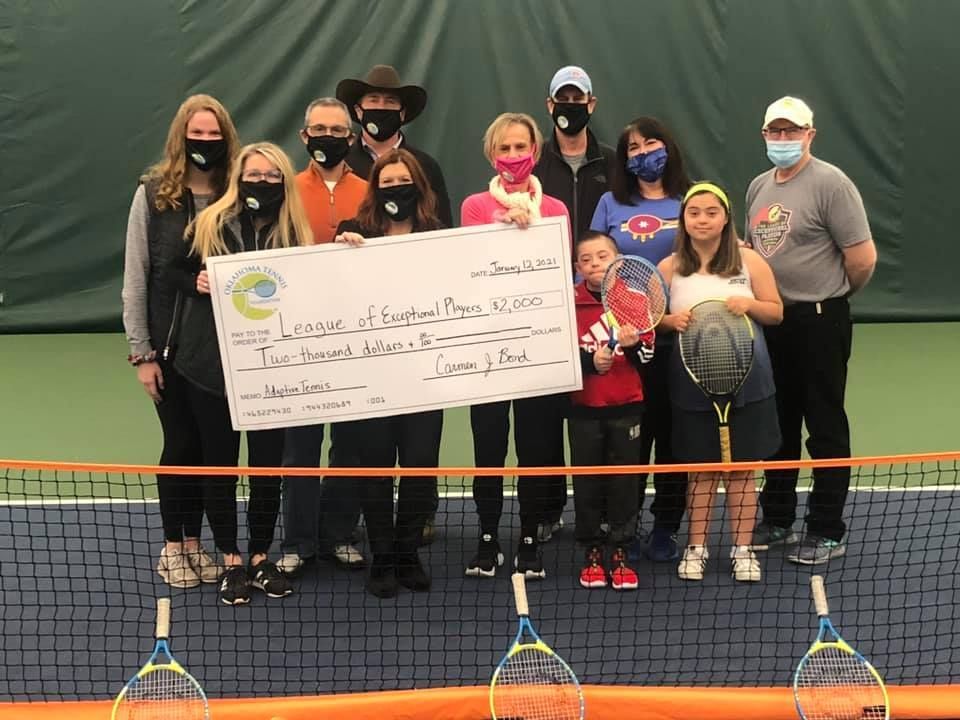 Tennis team poses indoors holding a large ceremonial check in front of a net