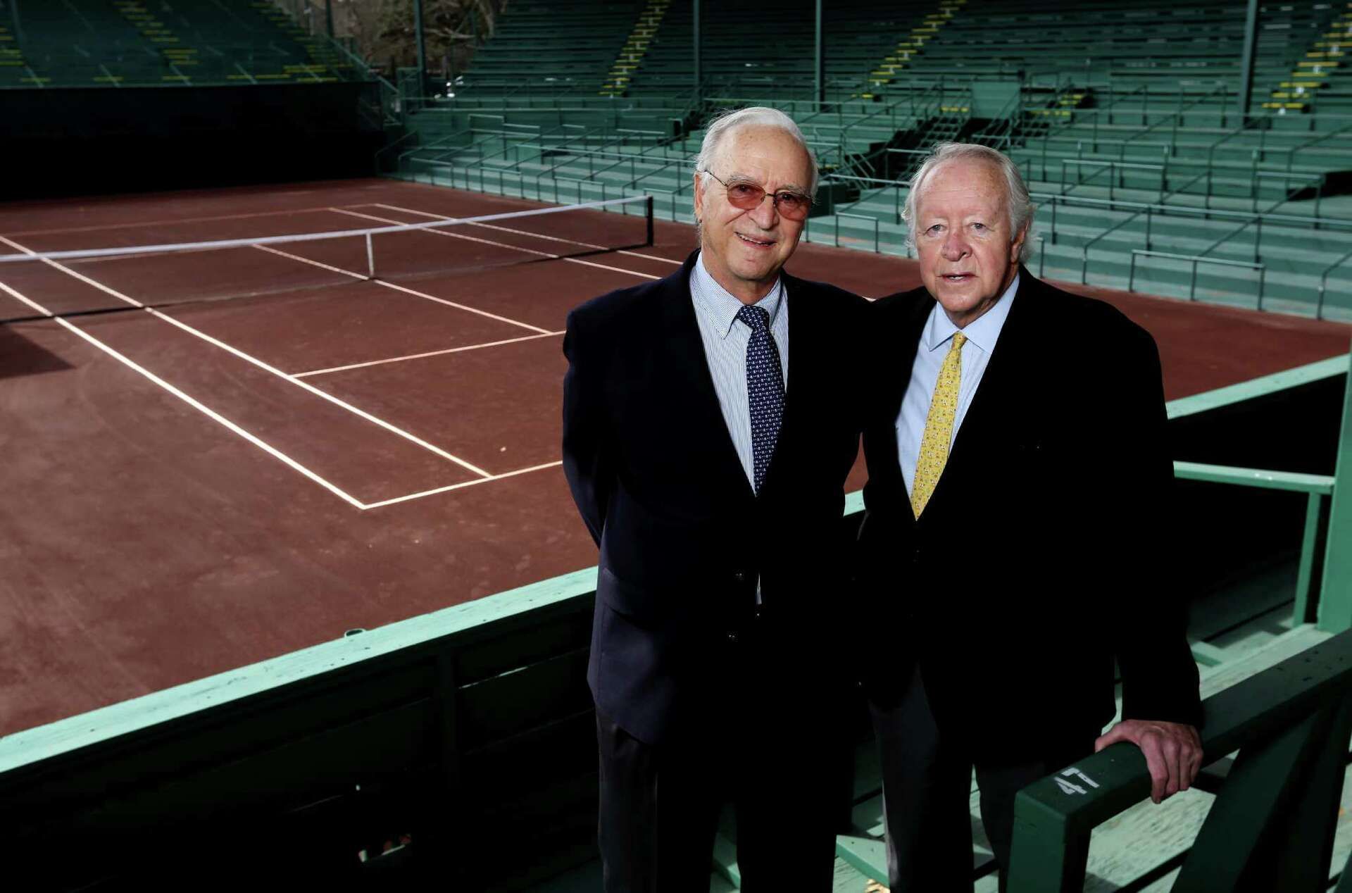 Two men in suits standing beside an indoor tennis court with green bleachers.