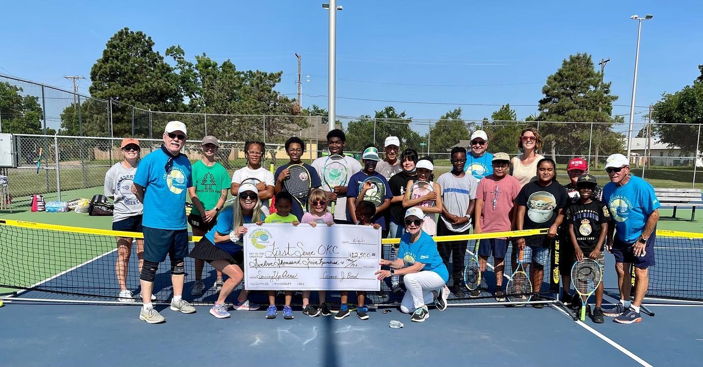 Group of people posing on a tennis court, holding a banner, with rackets and trophies in bright sunlight.