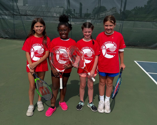Four children in red shirts pose with tennis rackets on an outdoor court.