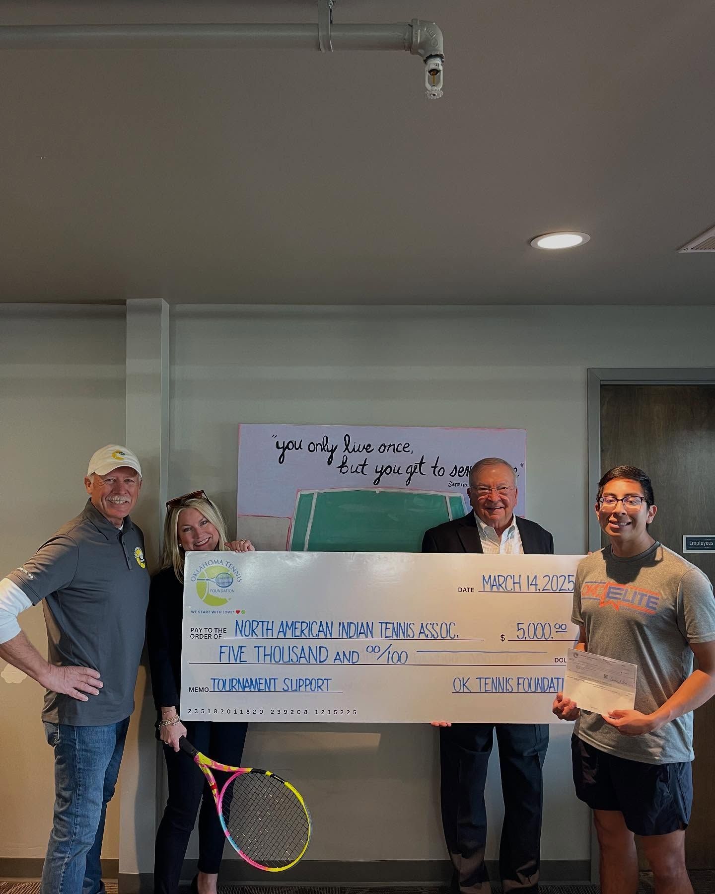 Three people indoors hold a large check and sign, with a tennis racket in the foreground.