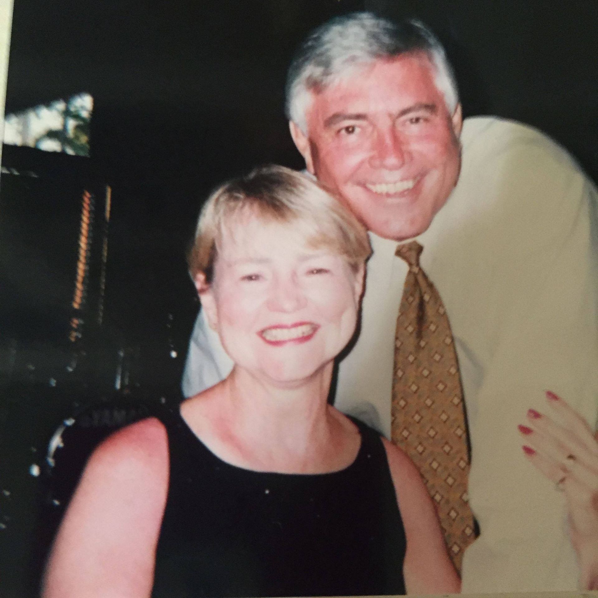 Two smiling people posing indoors, the woman in a black dress and the man in a tan shirt and tie.