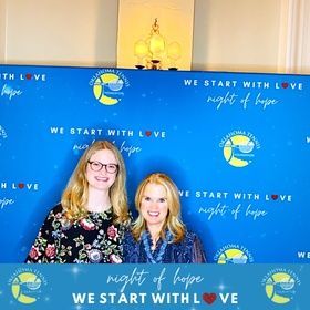 Two women posing before a blue “We Start with Love” and “Night of Hope” event backdrop