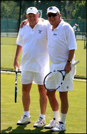 Two tennis players in white sportswear stand together on a grass court, holding racquets and smiling.