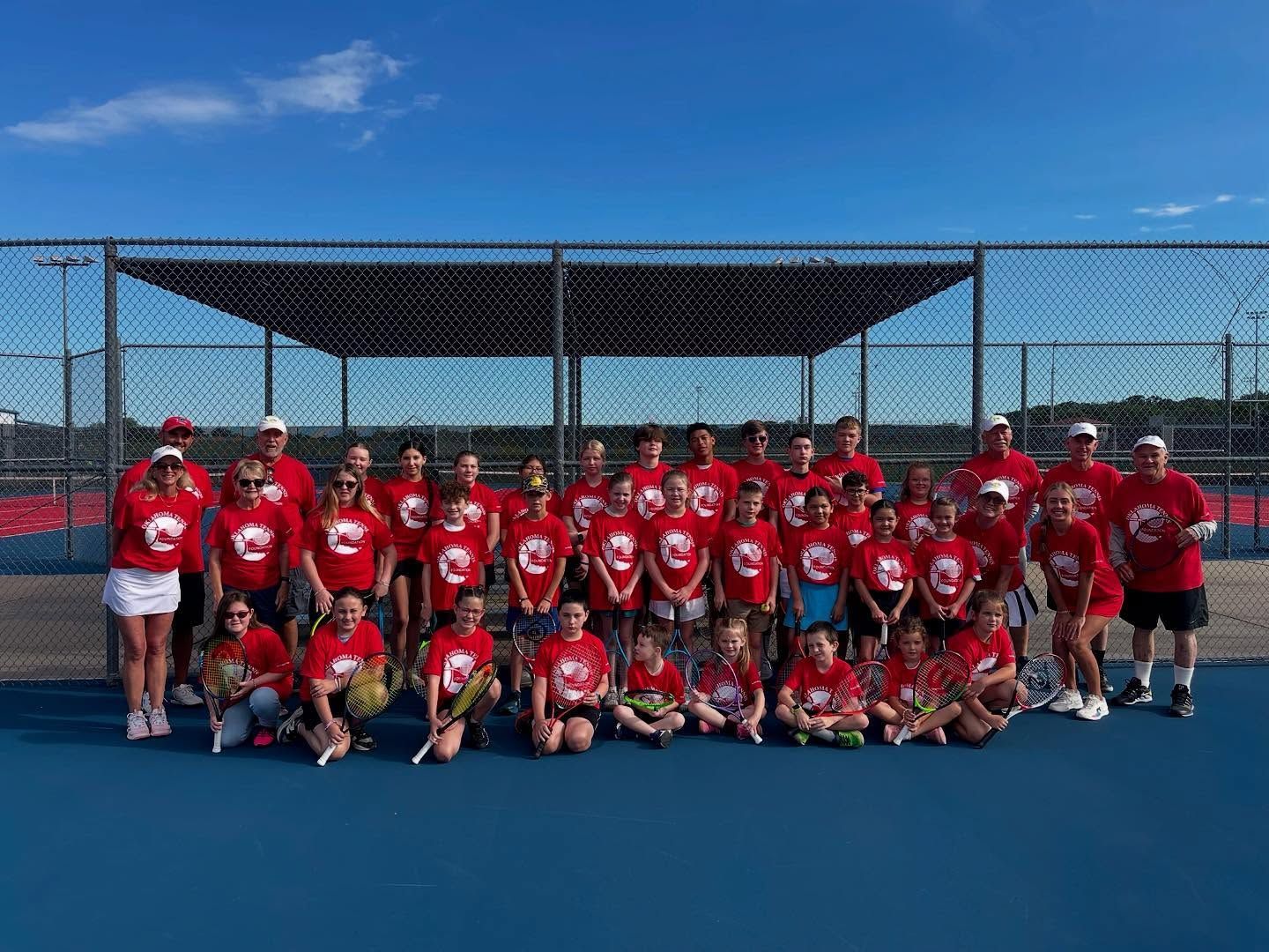 Youth sports team in red jerseys posing on a blue court under a covered fence, holding a trophy in front.