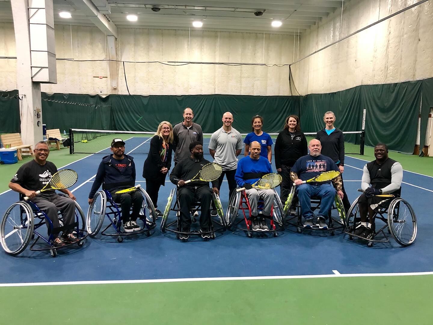 Group of people in wheelchairs posing indoors on a blue gym floor, smiling at the camera