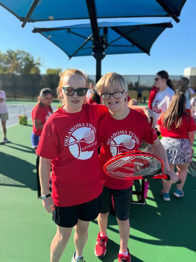 Two children in red shirts and sunglasses stand on a tennis court holding a racket under a blue umbrella.