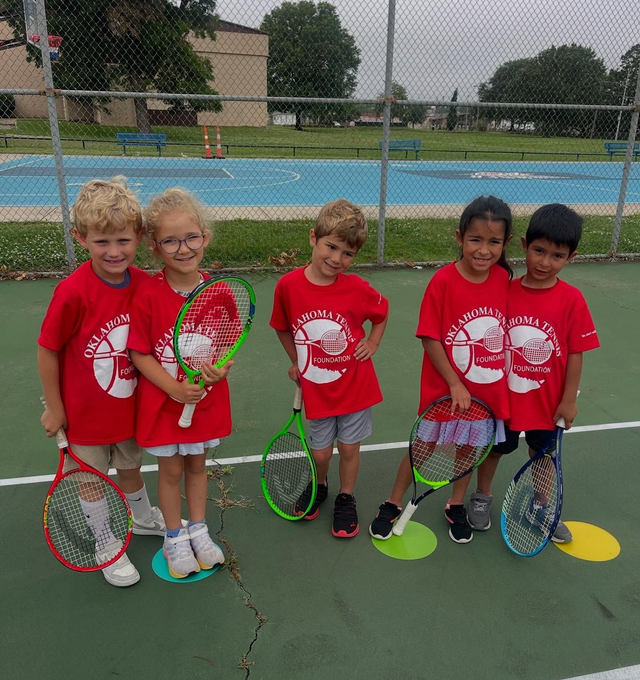 Five children in red shirts hold tennis rackets on a court with a pool in the background.