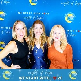 Three women posing at a Night of Hope event backdrop in blue, smiling and dressed in black, navy, and orange.