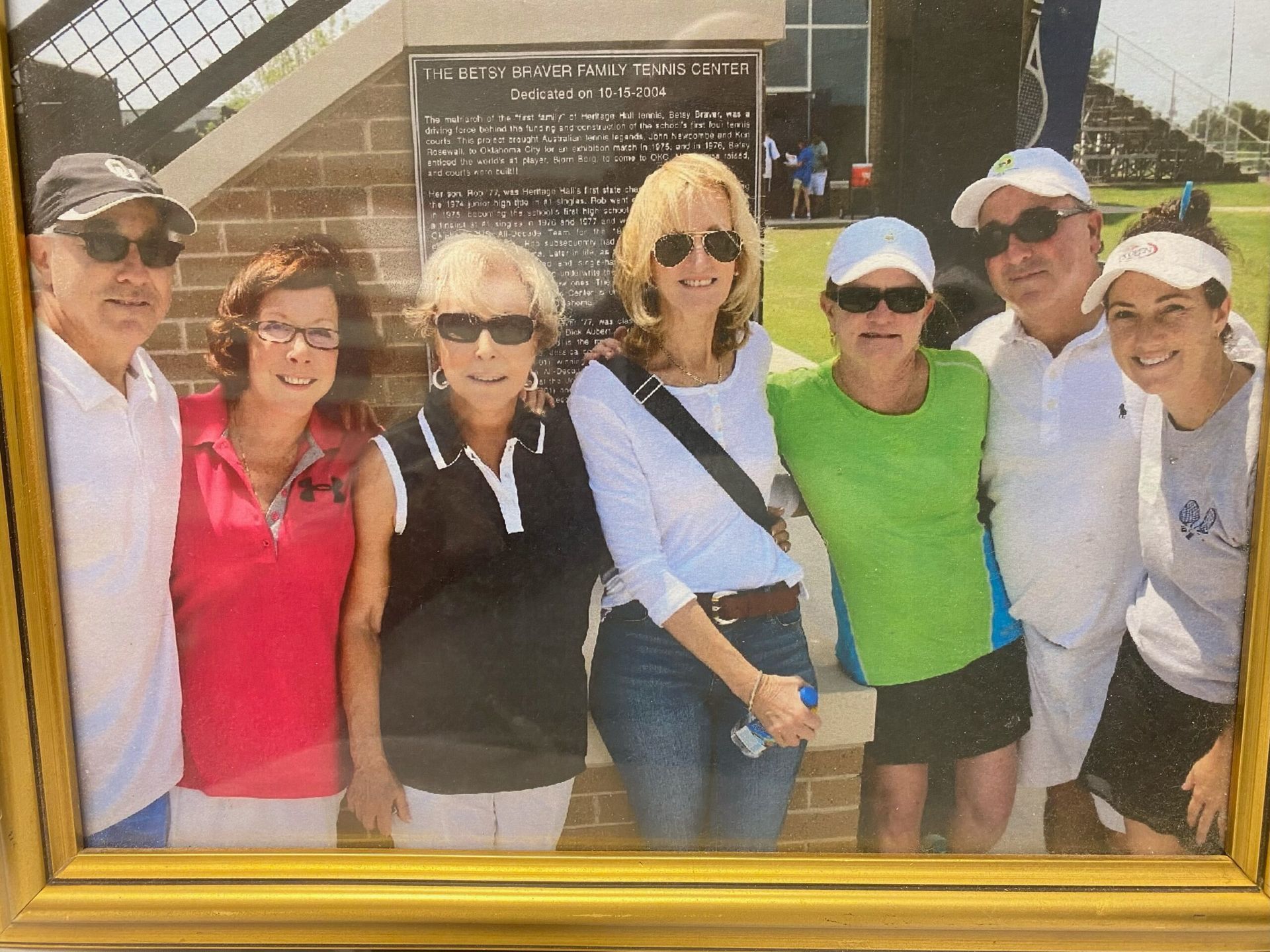 Six smiling adults posing in front of a framed plaque outdoors