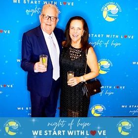 Two people posing at a blue “Night of Hope” event backdrop, holding drinks and smiling.
