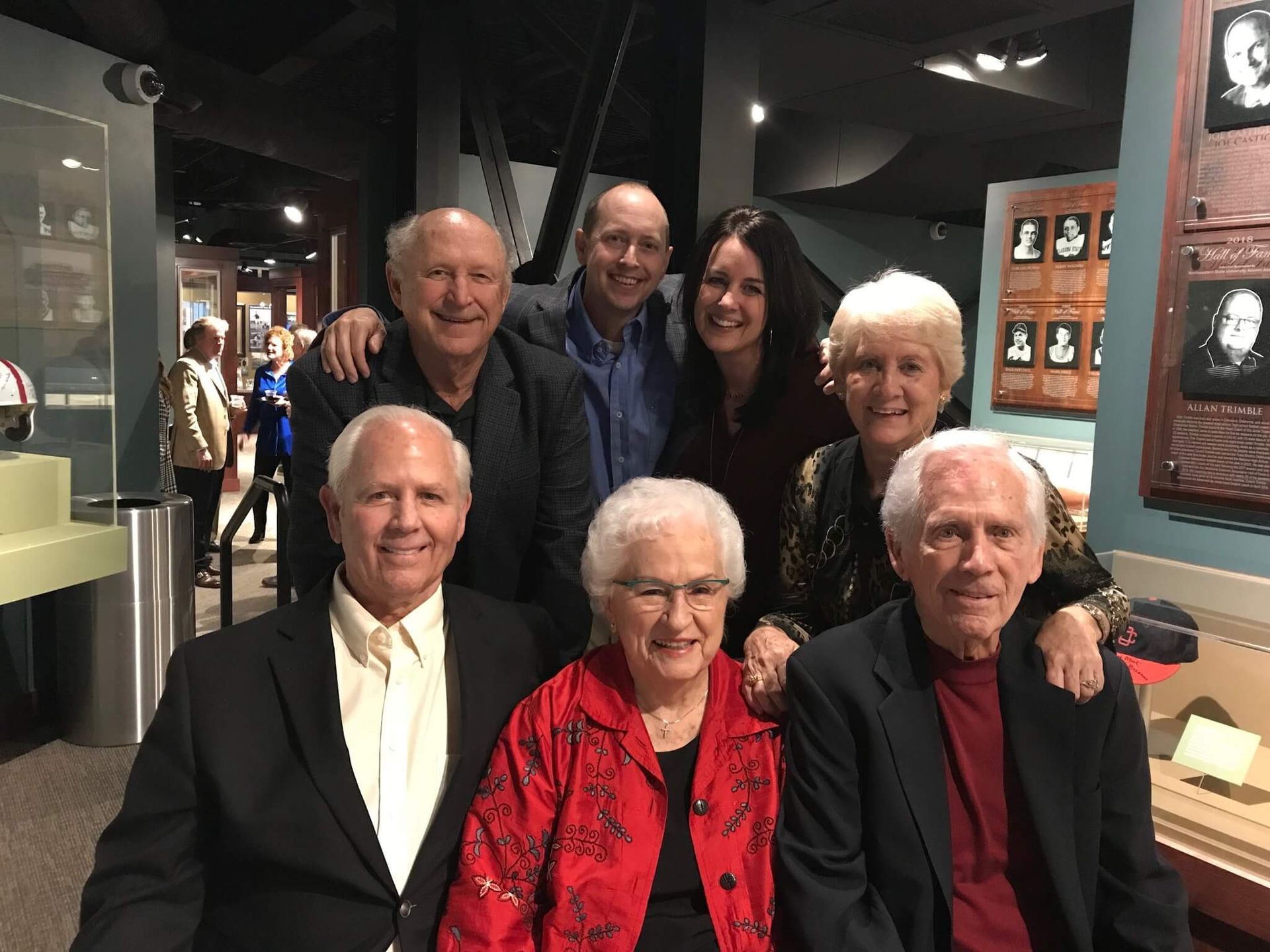 Six adults posing together in a museum-like gallery, smiling at the camera.
