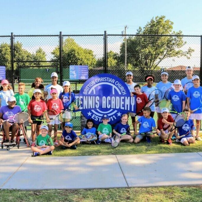 Group of children and adults posing outdoors with a blue “Tennis Academy” sign on a tennis court