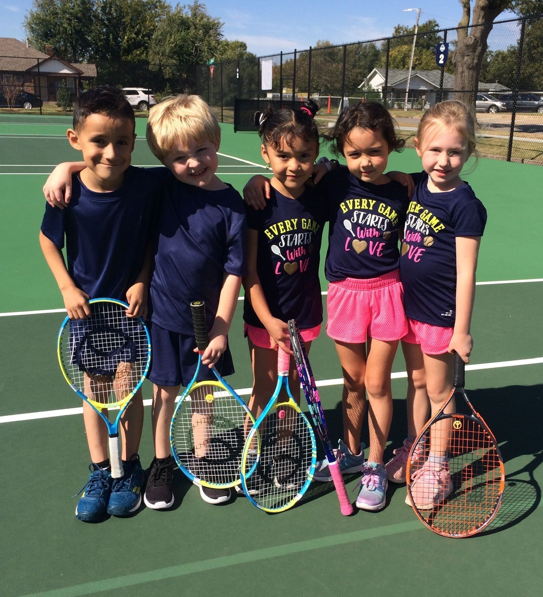 Five children holding tennis rackets stand together on an outdoor tennis court.
