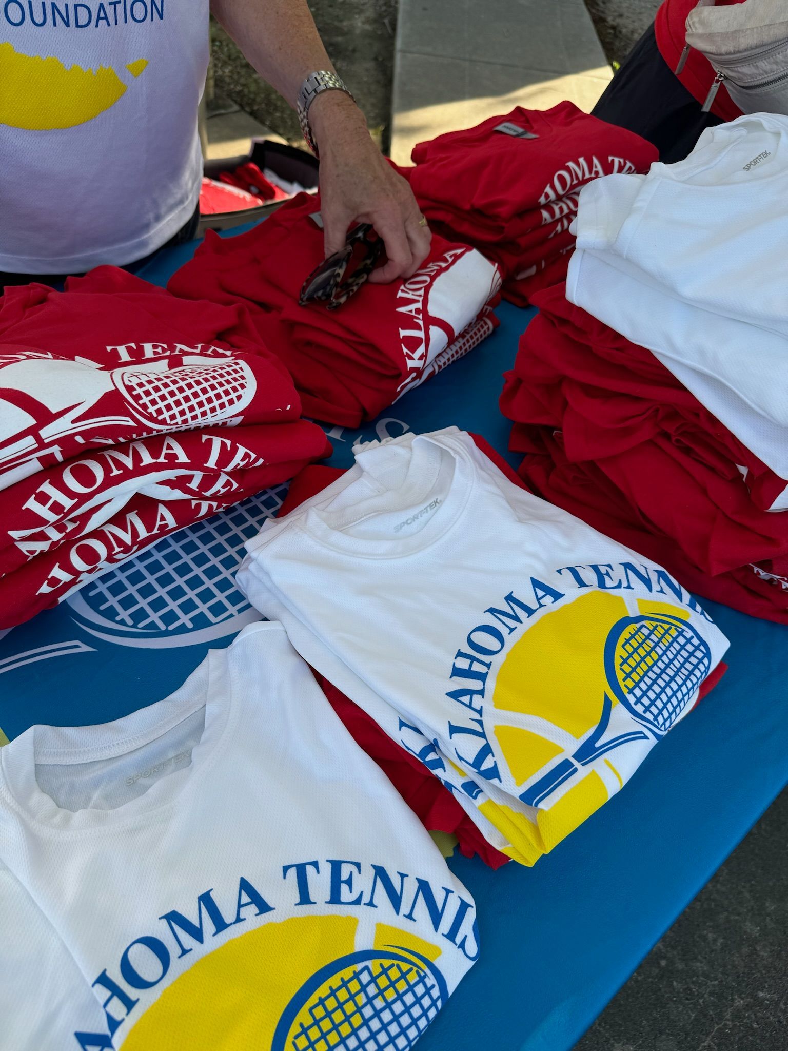 Stacked red and white Aloha Tennis T-shirts on a table, with a person arranging them