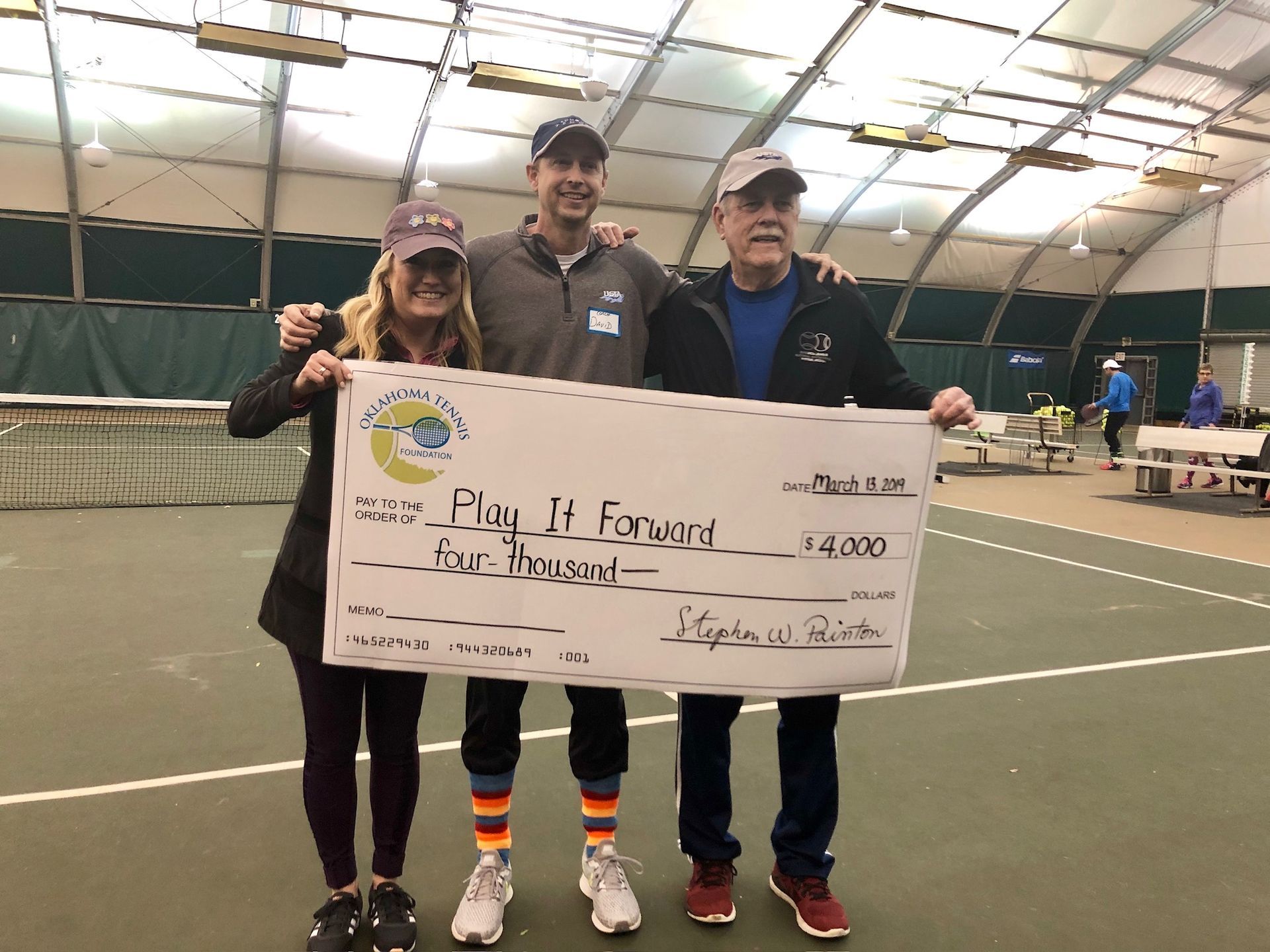 Three people holding a large check on an indoor tennis court.