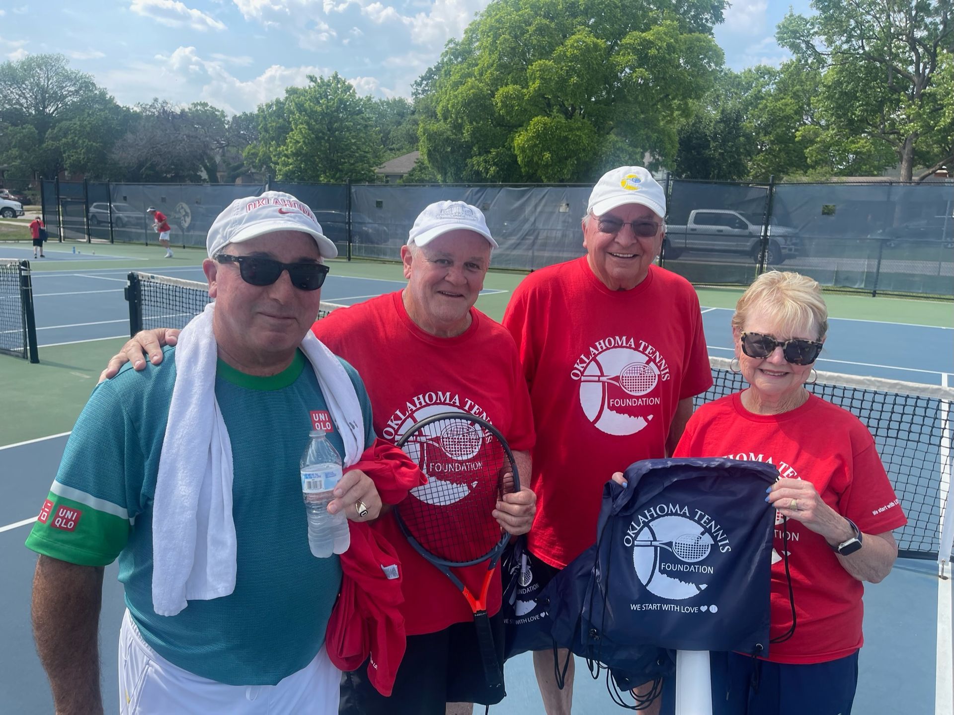 Four people in red shirts posing on a tennis court, two holding a navy event T-shirt.