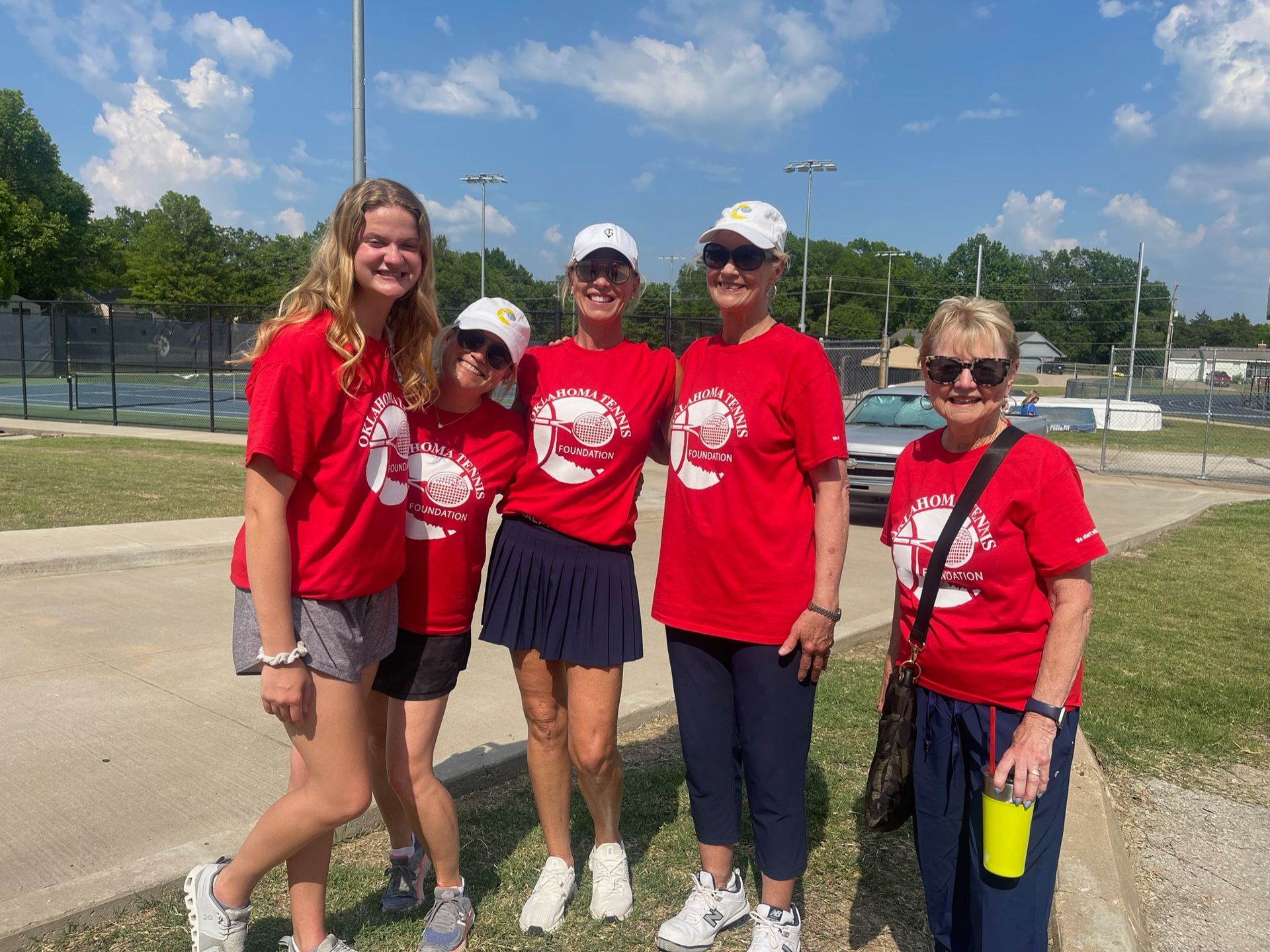 Five people in red shirts posing outdoors on a sunny day at a tennis court.