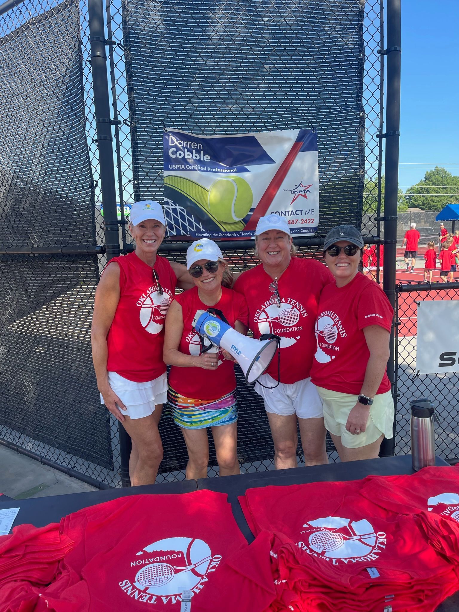 Four people posing at an outdoor event booth with red shirts, a banner, and display table