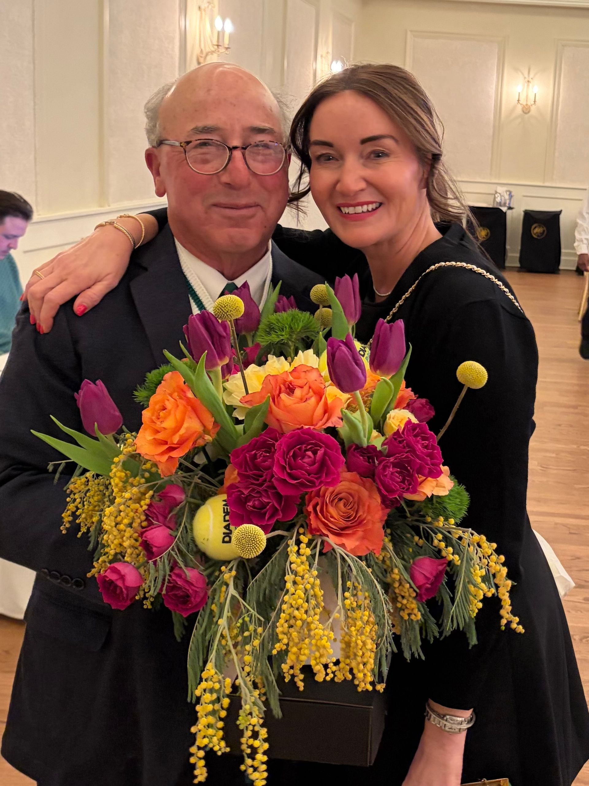 Two people smiling and posing with a large colorful flower bouquet indoors