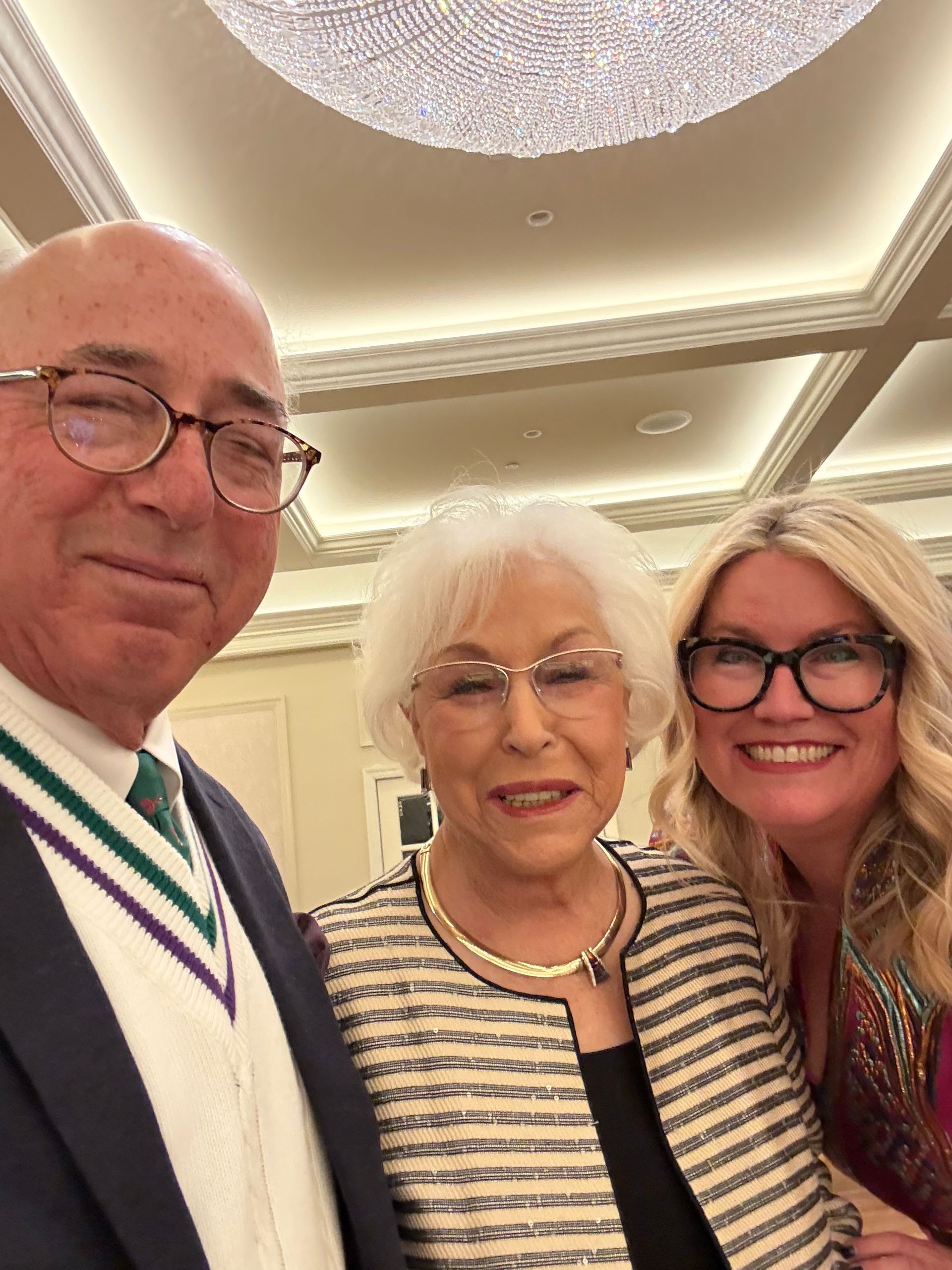 Three smiling people posing together in a bright ballroom, wearing formal attire.
