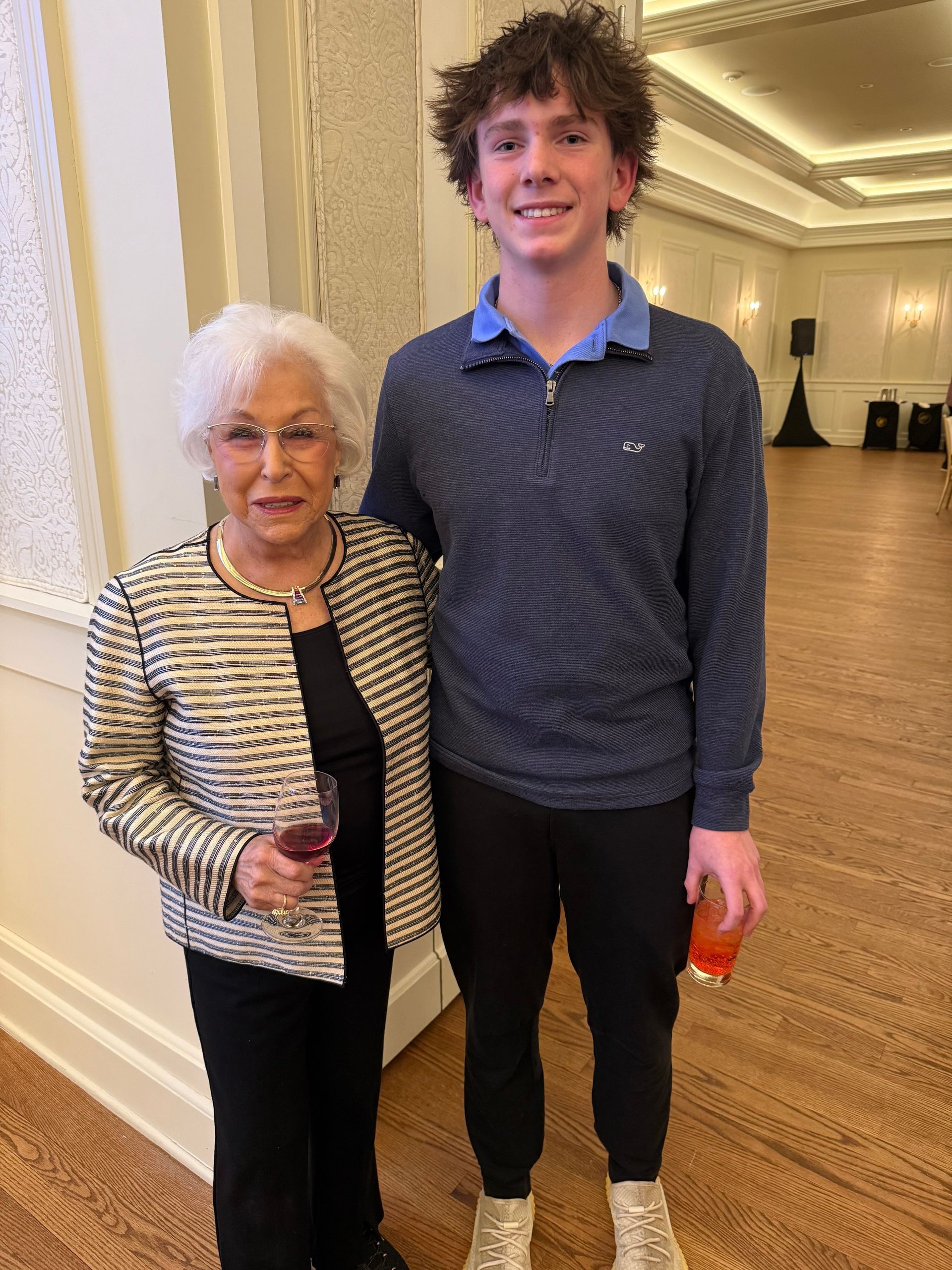 Two people standing together in a hallway, both smiling, one holding a drink.