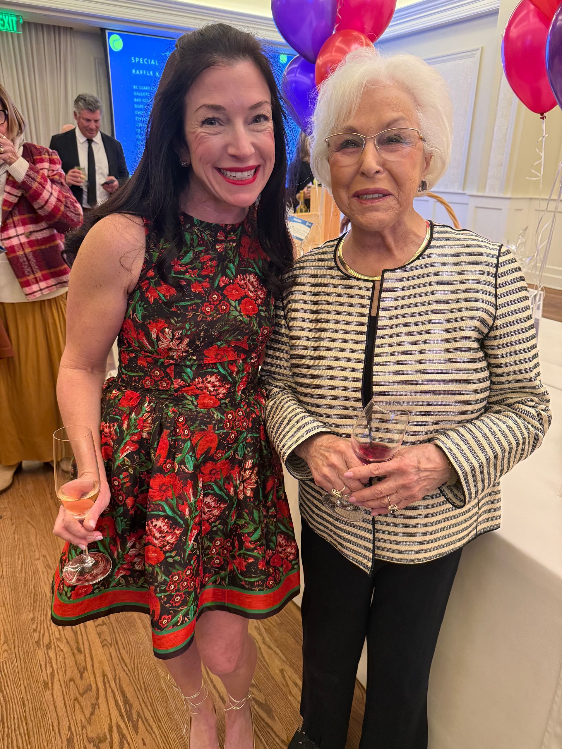 Two women smiling indoors at a festive event, wearing colorful outfits and standing by balloons.