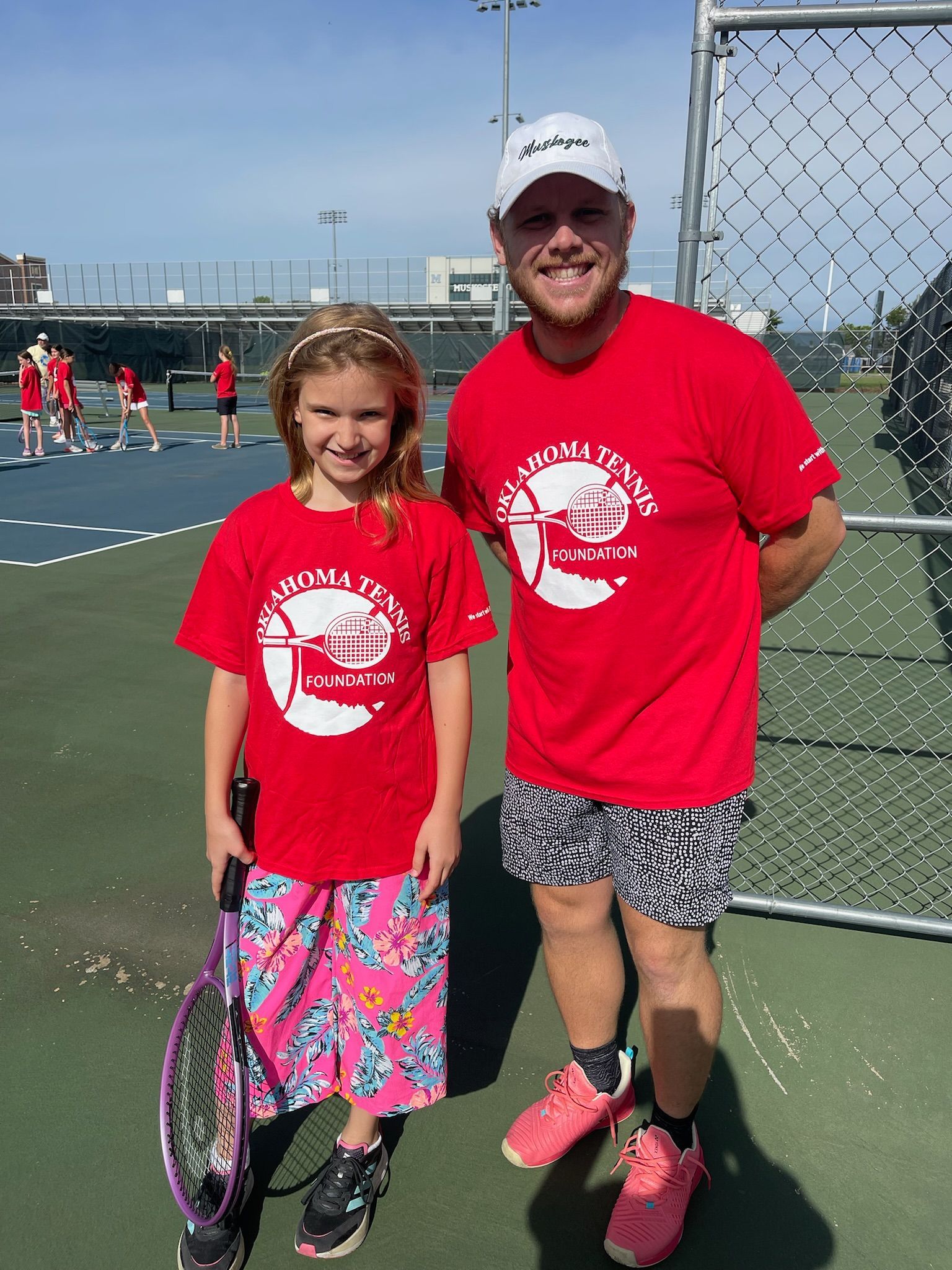 Two tennis players in red shirts stand on an outdoor court holding rackets, smiling at the camera.