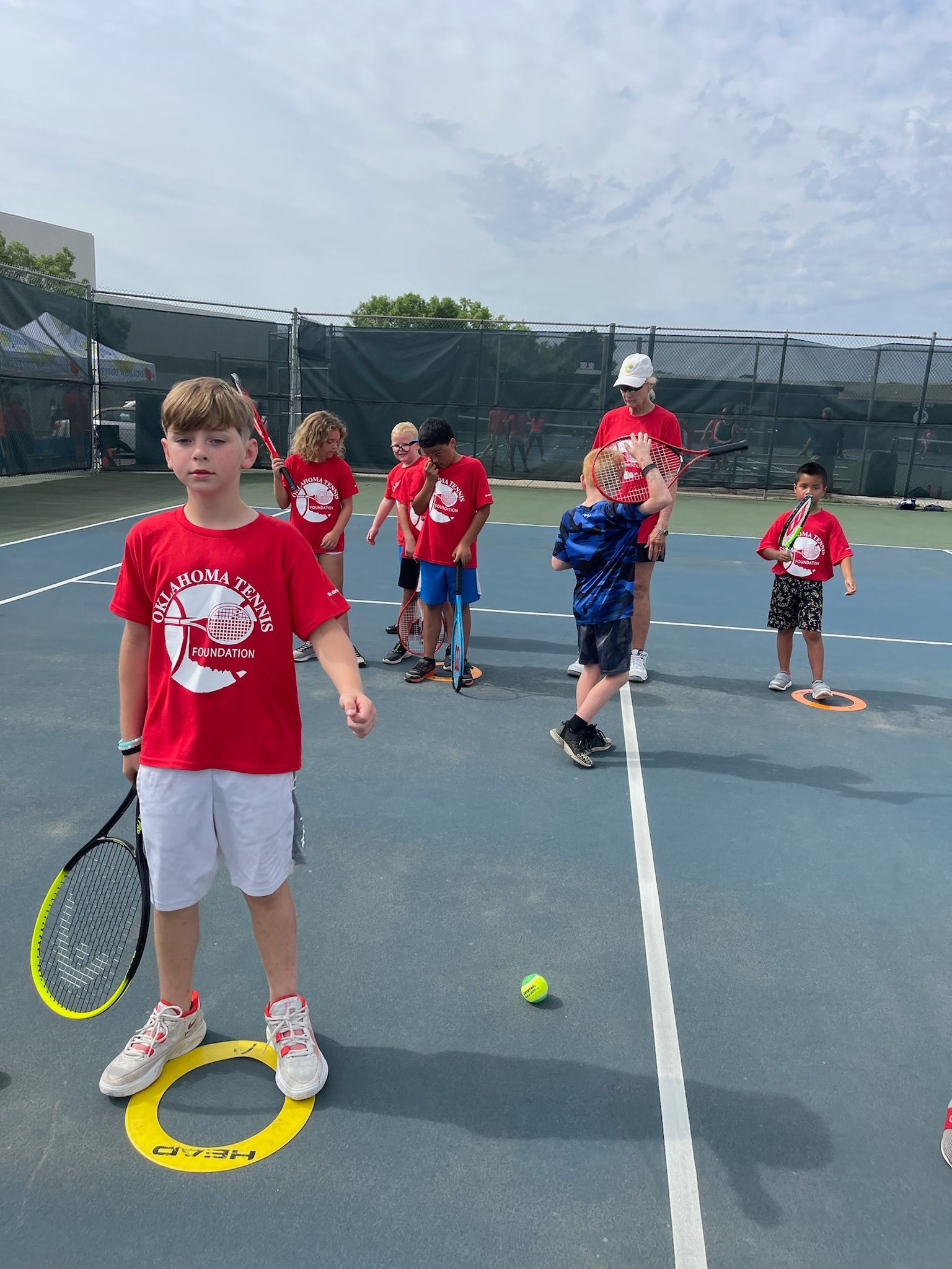 Children holding tennis rackets on an outdoor court, with tennis balls scattered on the ground.