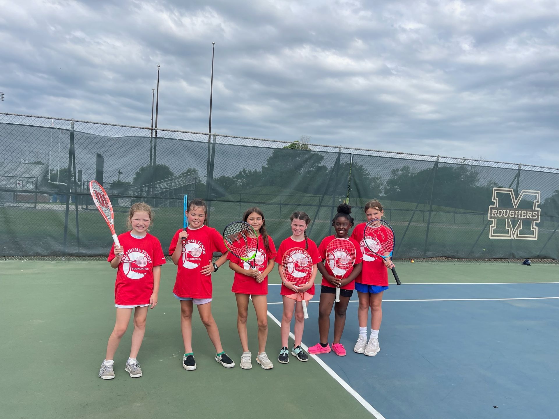 Six youths in red shirts stand with tennis rackets on a court, posing near a netted fence.