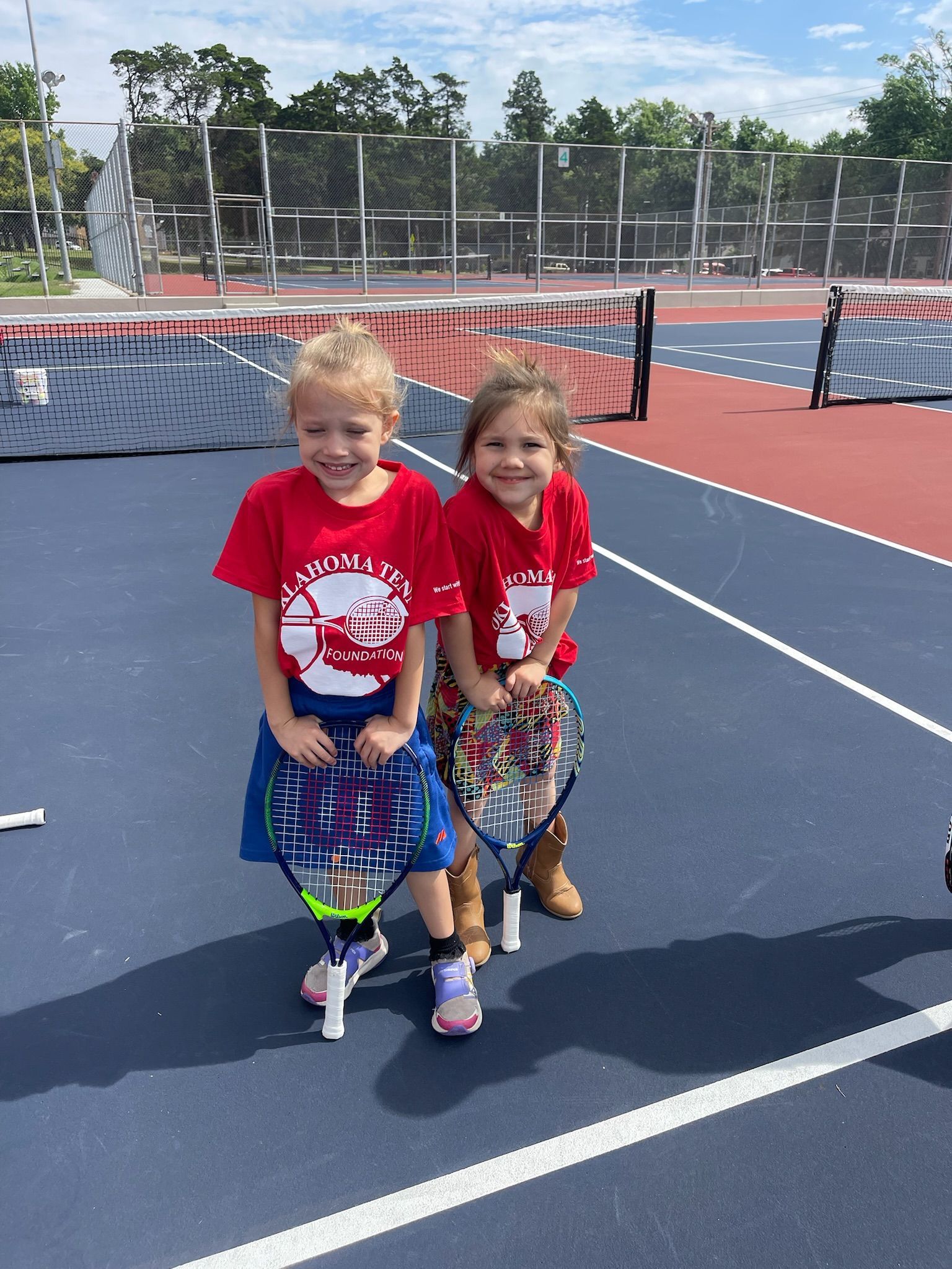 Two children in red shirts stand on a tennis court, smiling and holding bags near the net.