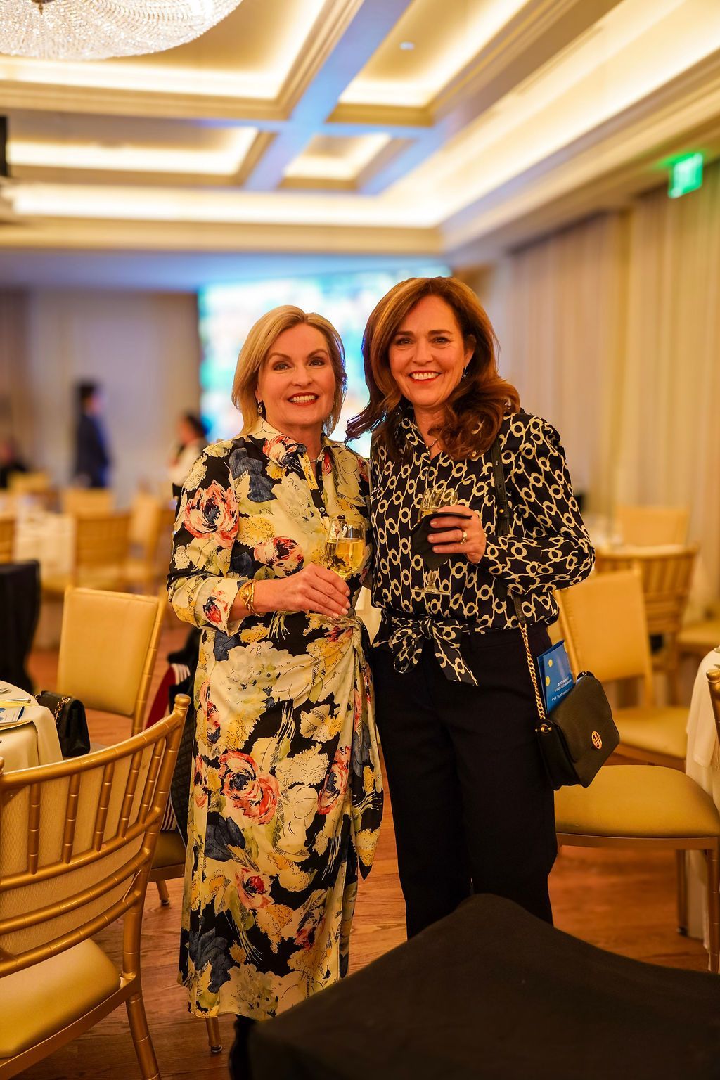 Two women smiling and posing with drinks at a warmly lit indoor event venue