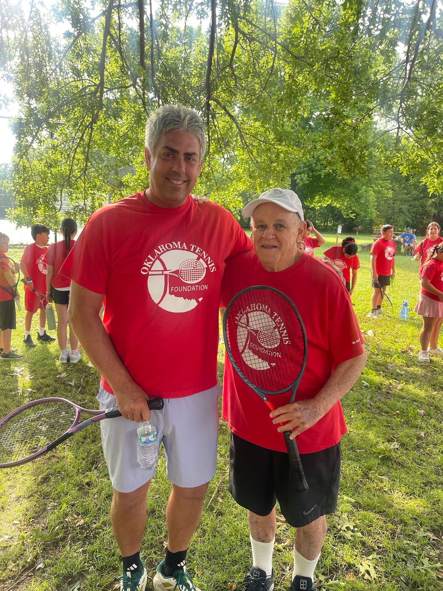 Two people in red shirts pose on a grassy court holding tennis rackets under trees.