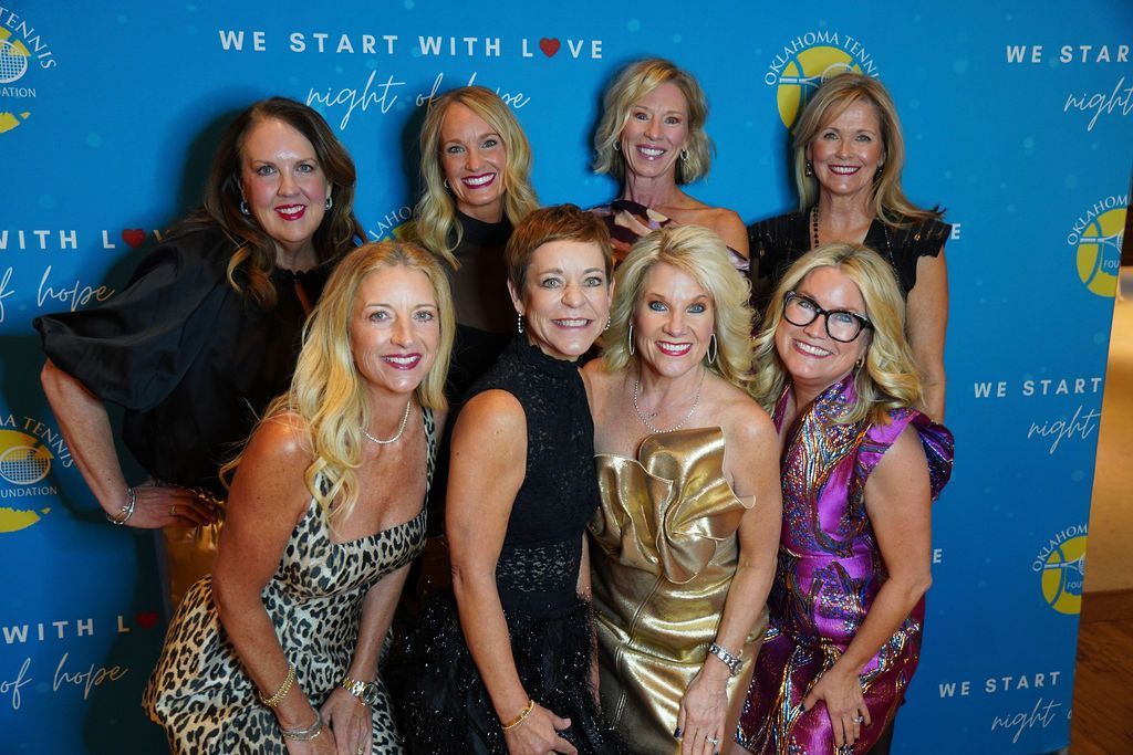 Seven women posing on a blue event backdrop with the words “We Start With Love” repeated behind them