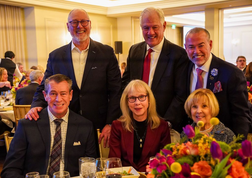 Five people pose smiling at a banquet table with flowers in a warmly lit dining room.